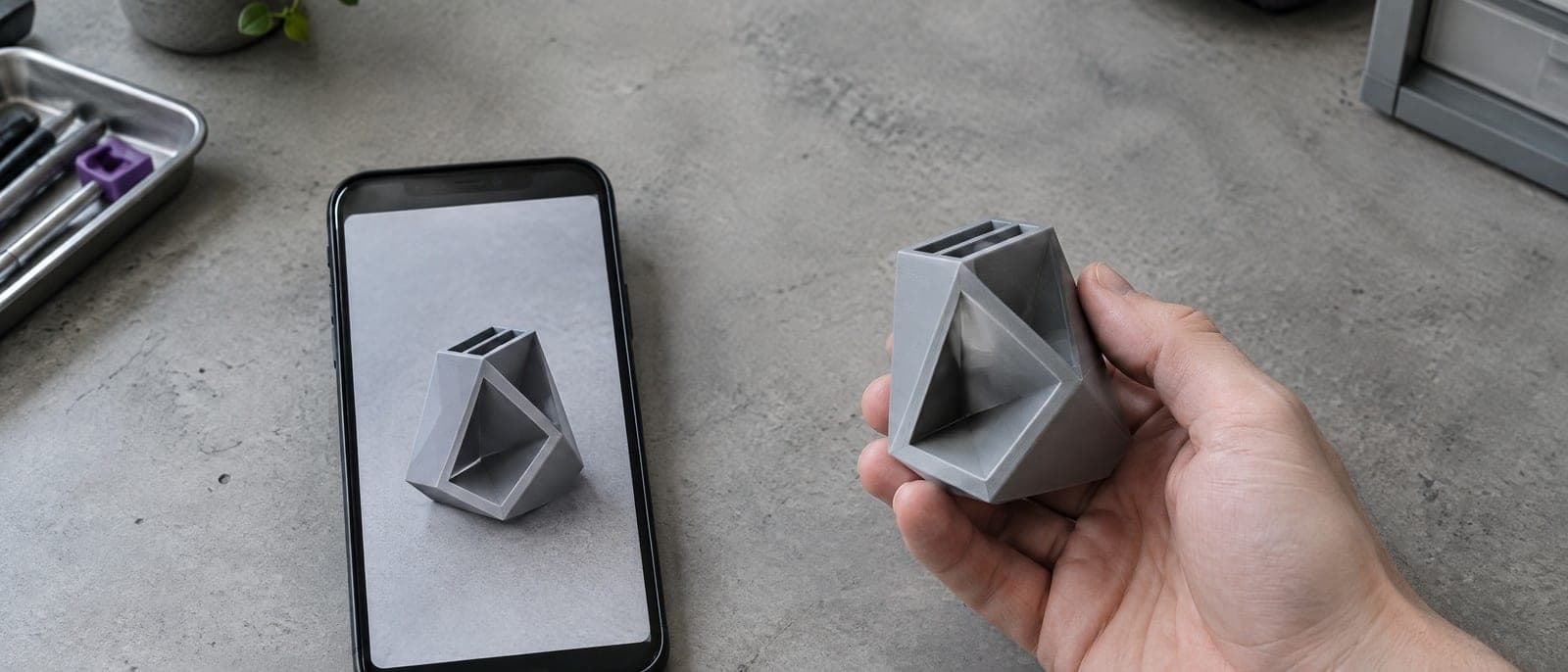 Een telefoon met een foto van een hoekig grijs houdertje, en daarnaast een hand die hetzelfde voorwerp als 3D-print vasthoudt.
