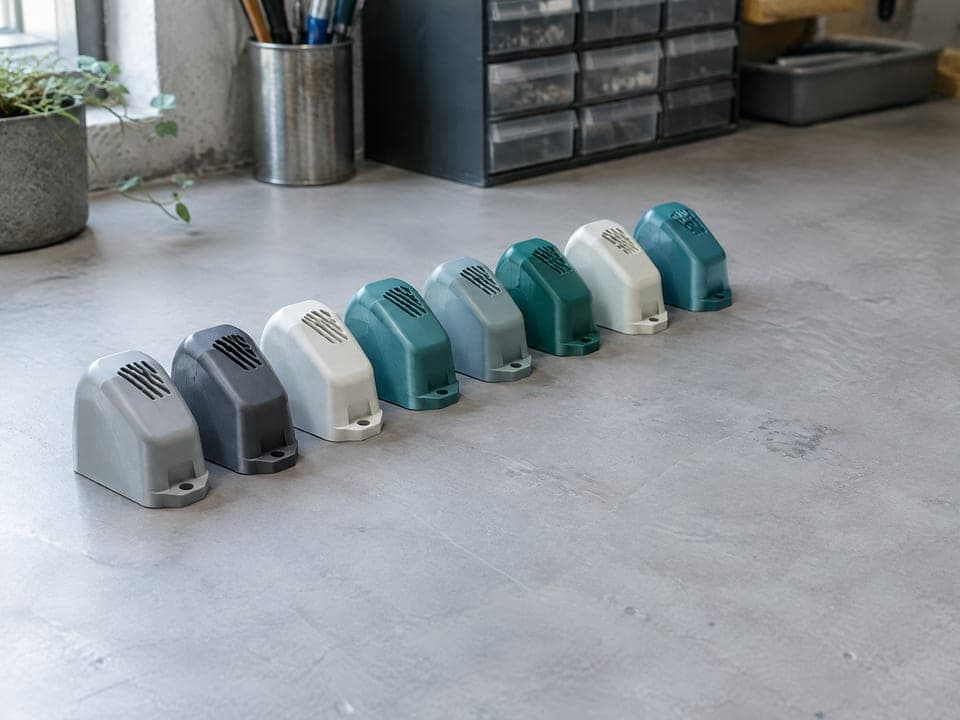 Een rij van acht identieke kleine behuizingen met ventilatiesleuven, elk in één effen kleur: grijs, antraciet, wit en blauwgroen.